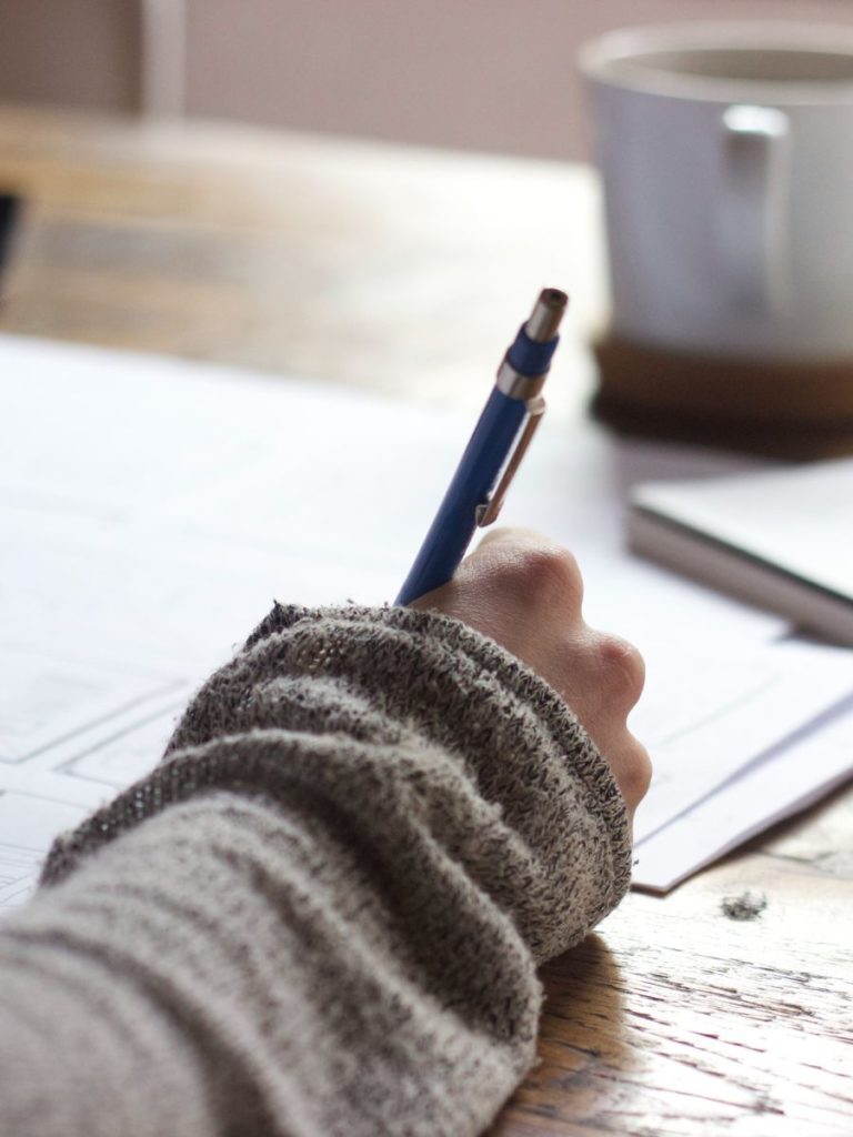 A woman's arm in a sleeve is resting on a desk. She is holding a pen and writing in a notebook.