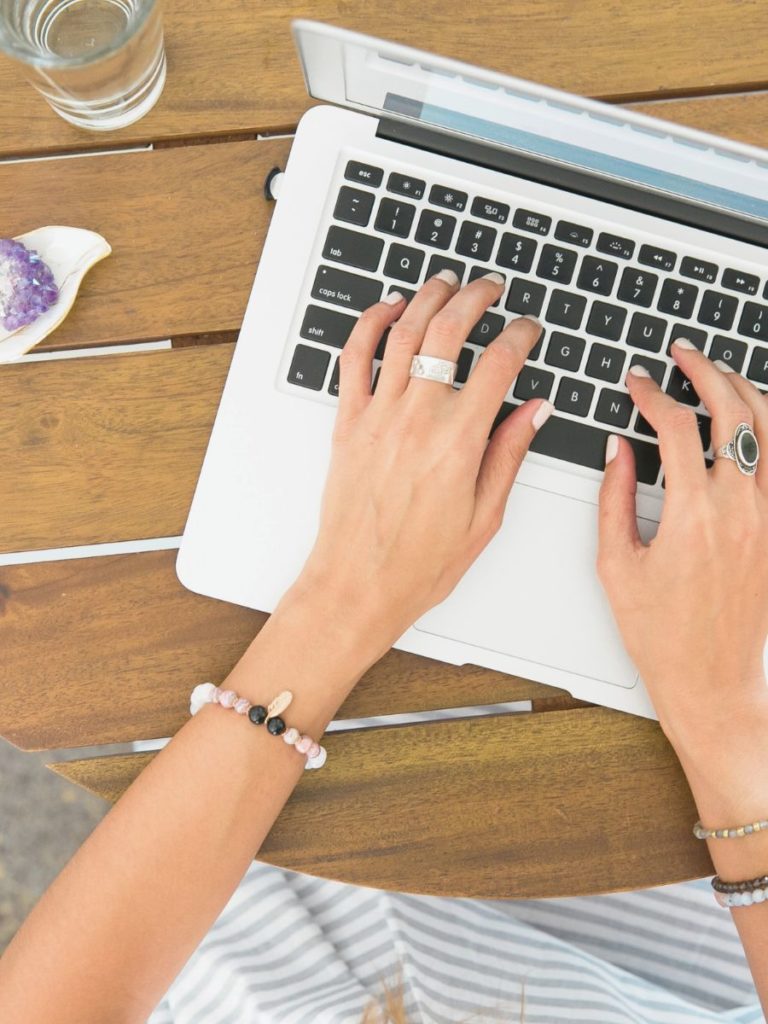 An overhead photo of a woman typing on a laptop, wearing bracelets and rings